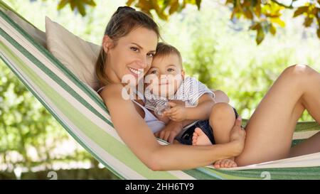 Lächelnde und glückliche Mutter umarmt ihren kleinen Sohn und legt sich entspannt in der Hängematte, im Hausgarten an einem schönen und sonnigen Sommertag, freudig Stockfoto