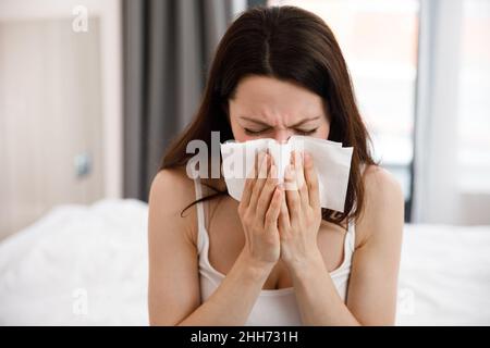 Eine kranke junge Frau bedeckte ihre Nase. Sie hat Fieber, sie hat eine Erkältung, niest in eine Serviette, sitzt auf dem Bett. Eine kranke allergische Frau mit Allergie Stockfoto