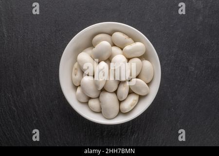 Getrocknete Körner aus biologischen weißen Bohnen in einer Keramikschale auf einer Schiefertafel, Draufsicht. Stockfoto