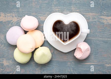 Runde mehrfarbige Marschmeichs in einem Stapel, eine abgebissene Hälfte eines Marschmeiches und eine weiße herzförmige Tasse mit schwarzem dampfendem heißen Kaffee auf einem weißen Stockfoto