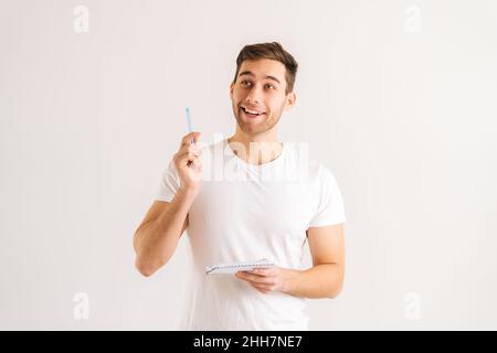 Studio-Porträt eines enthusiastischen jungen Mannes, der im Copybook schreibt und eureka-Geste auf weißem, isoliertem Hintergrund hochhebt. Stockfoto