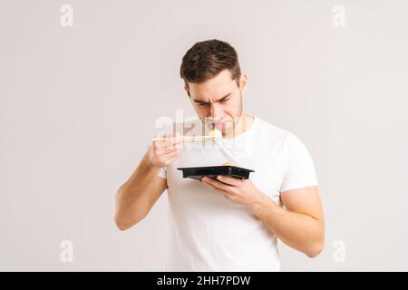 Porträt eines angewiderten jungen Mannes, der Sushi mit Essstäbchen und Schnüffeln hält, mit einem Abscheu im Gesicht auf weißem, isoliertem Hintergrund. Stockfoto
