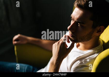 Nahaufnahme aus einem hohen Winkel Gesicht eines wirklich schönen Mannes, der das Kinn nachdenklich berührt und auf einem gelben Stuhl auf dunklem Hintergrund sitzt Stockfoto