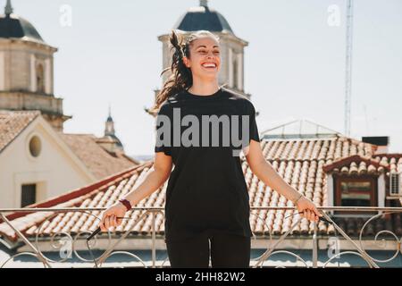 Sportliche Frau in stilvoller Sportkleidung, die am sonnigen Tag beim Training auf dem Dach des Gebäudes die Seilsprungübung macht. Stockfoto