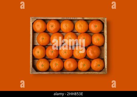 Holzkiste mit reifen Mandarinen auf orangefarbenem Hintergrund. Clementine Mandarinen in Holzkiste. Blick von oben. Geeignet für nahtlose Mustererstellung. Stockfoto