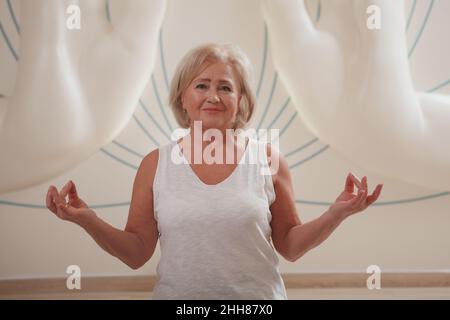 Reizende ältere Frau, die lächelt und Handmudras macht, während sie meditiert Stockfoto