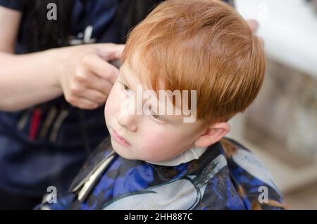 Die Hände des Friseurs schneiden dem Jungen die Haare mit einem Trimmer. Rothaariger Junge im Friseurladen. Stockfoto