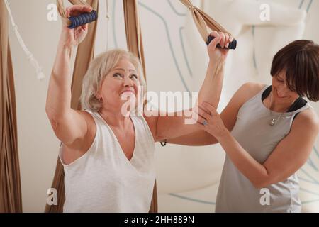 Fröhliche ältere Frau lächelt, während sie mit einem persönlichen Trainer in der Hängematte Luftyogas praktiziert Stockfoto