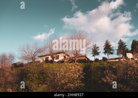 Dorfhaus auf einem Hügel mit Bäumen unter blauem und bewölktem Himmel, schöne Landschaft eines Dorfhauses auf grüner ländlicher Umgebung in Birgi, Odemis, Izmir. Stockfoto