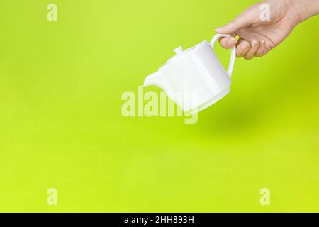 Frau Hand gießt Tee aus weißer Keramik Teekannen vor hellgrünem Hintergrund. Tee brauen und trinken. Stockfoto