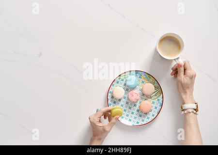 Draufsicht auf bunte Kuchenmakronen auf einem Teller und weibliche Hand mit einer Tasse Kaffee Stockfoto