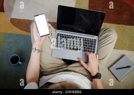 Draufsicht auf jungen Mann mit Laptop und Smartphone, während er auf dem Boden in einem modernen Apartment sitzt Stockfoto