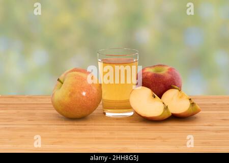 Ein Glas Apfelsaft mit Äpfeln und Apfelscheiben auf einem hölzernen Schneidebrett, isoliert auf einem grünen Bokeh-Hintergrund Stockfoto
