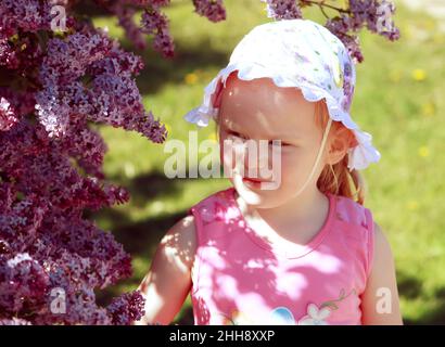 Schönes kleines blondes Mädchen mit panama auf dem Kopf, das im Frühlingsgarten mit blühendem Busch aus purpurem Flieder spielt. Stockfoto