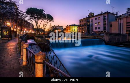 Treviso, die San Martino Brücke bei Sonnenuntergang. Auf der rechten Seite ein Wasserfall auf dem Fluss Sile, auf der linken Seite ein Spaziergang von Straßenlaternen beleuchtet Stockfoto