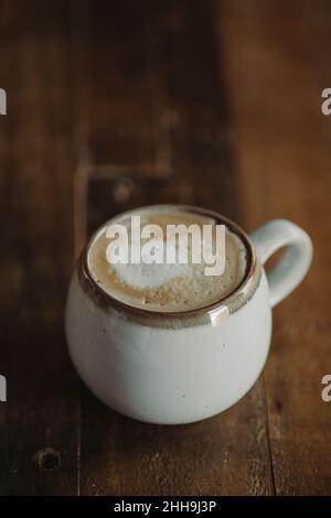 Isolierter Cappuccino im weißen Becher am Tisch aus dunklem Holz. Moody Coffee Shop Lifestyle. Kaffee flach Lay mit Copy Space. Cortado. Flach Weiß. Stockfoto