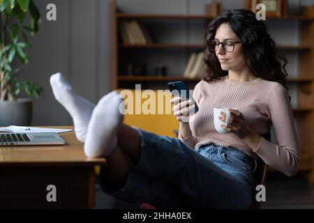 Entspannte junge Freiberuflerin, die sich mit Mobiltelefon und Kaffeetasse am Schreibtisch ausruhen und freiberuflich arbeiten kann Stockfoto