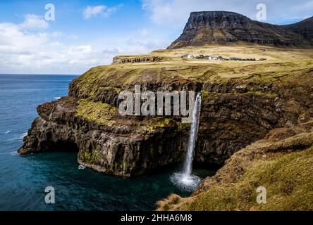 Ein Wasserfall stürzt sich über eine dramatische Felswand in eine wilde weiße Spritzzone. Ein Dorf liegt ruhig darüber, eingebettet in hellgrünes Gras. Stockfoto