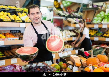 Junger Mann Verkäufer hält die Hälfte der Wassermelone in den Händen im Obstladen Stockfoto