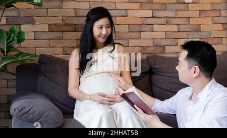 Erwachsener Mann liest ein Buch und berührt den Bauch seiner Schwangeren, während er auf einem bequemen Sofa sitzt Stockfoto