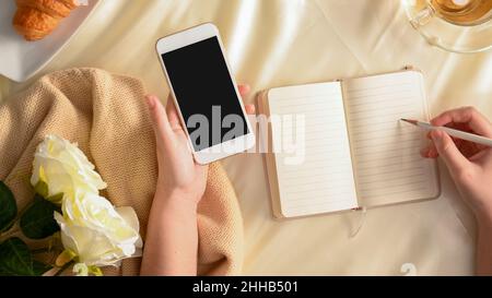 Flat Lay, Büroschreibtischkonzept für Frauen. Frau notiert sich ihr Buch und hält ein Smartphone über den Arbeitstisch. Stockfoto