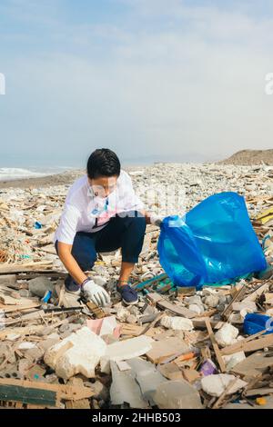 Vertikales Bild einer lateinischen Freiwilligen, die Müll an einem verschmutzten Strand aufsammelt. Stockfoto