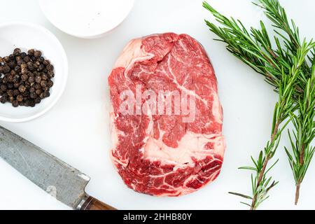 Nahaufnahme eines rohen Rib-Eye-Steakes, Pfefferkörner, Salz und ein paar Zweige frischen Rosmarins auf einer weißen Oberfläche. Lebensmittelkonzept. Ansicht von oben. Stockfoto