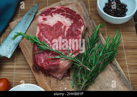 Ein Auge für rohes Rindfleisch auf einem Holzschneidebrett neben einem Feldmesser und einigen Rosmarin- und Pfefferkörnern. Lebensmittelkonzept. Stockfoto