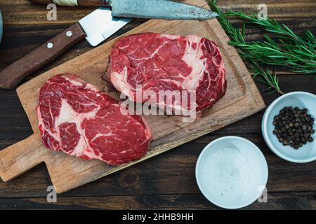 Zwei rohe Rippenaugen auf einem rustikalen Holzschneidebrett und Pfefferkörnern, Salz und ein paar Zweige frischen Rosmarins. Lebensmittelkonzept. Ansicht von oben. Stockfoto