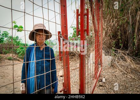 Eine ältere Frau schaut durch den roten Zaun, der die Goldmine von Tungkum umgibt. Seit über einem Jahrzehnt kämpft die Khon Rak Ban Kerd Gruppe aus einem kleinen ländlichen Dorf in der Provinz Loei in Thailand gegen die Goldmine Tungkum. Die Gruppe besteht aus angesehenen - und größtenteils Analphabeten - älteren Frauen, die ihren Aktivismus durch den Anbau und die Verarbeitung von Bio-Baumwolle finanziert haben. Die Landwirtschaft ist die Haupteinnahmequelle für die Mehrheit der Einwohner von Loei; Ressourcen wie Wasser und Land haben einen großen Einfluss auf ihre Lebensgrundlage. Wenn die lokale Gemeinschaft Symptome berichtet, die mit Blut übereinstimmen Stockfoto