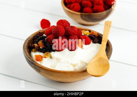 Holzschüssel mit griechischer Joghurt-Schlagsahne mit Himbeeren und getrockneten Früchten, Nahaufnahme auf weißem Hintergrund Stockfoto