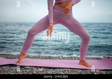 Gut aussehende Frau mittleren Alters mit schwarzen Haaren, Fitnesstrainerin in Leggings und Tops beim Stretching und Pilates auf einer Yogamatte in der Nähe des Meeres. Weiblich Stockfoto