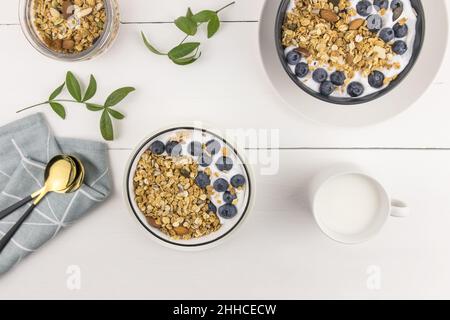 Draufsicht auf drei Schalen mit gebackenem Müsli und frischer Milch, Heidelbeeren und Nüssen. Leckeres, gesundes Frühstück. Weißer Holzhintergrund Stockfoto