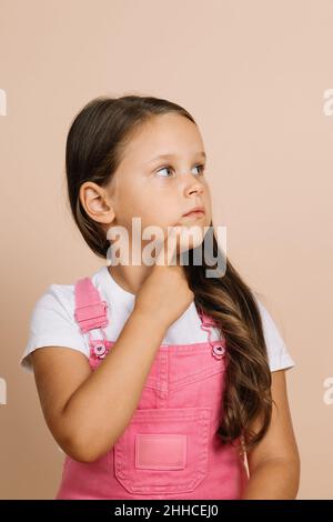 Porträt eines Kindes mit leuchtenden, hellen Augen, die wegschauen, zeigen Sie mit dem Finger auf das Kinn im Gesicht mit nachdenklicher Optik und tragen Sie einen leuchtend rosa Jumpsuit und einen weißen T Stockfoto