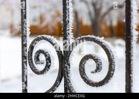 Mittlerer Teil des schwarzen schmiedeeisernen Zauns mit verschwommenem Holzhaus und Bäumen im Hintergrund. Schnee bedeckt Zaun und macht es noch fantastischer aussehen Stockfoto