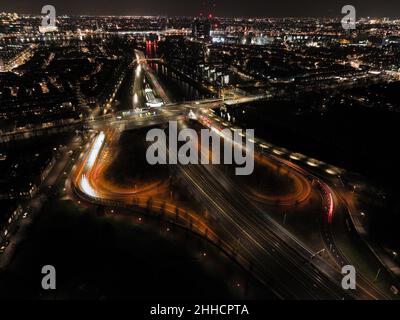 Amsterdam in der Nacht, holländische Infrastruktur Straßenkreuzung, Zug, Straße, Strecke, U-Bahn und Fußgänger. Luftdrohnenansicht. Stockfoto