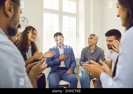 Eine Gruppe verschiedener Personen sitzt in einem Kreis und applaudiert ihrem männlichen Kollegen bei einem Teamtreffen. Stockfoto