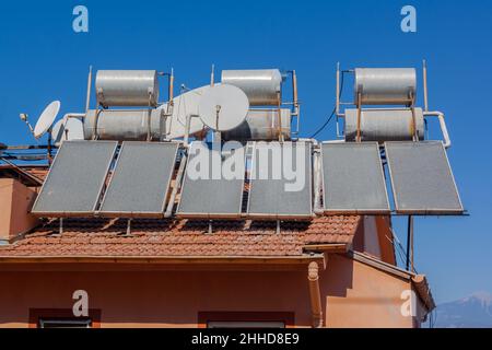 Viele Solarwasserheizsysteme und Satellitenschüsseln auf dem Dach des Hauses. Viele große Wassertanks auf Fliesendach. Stockfoto