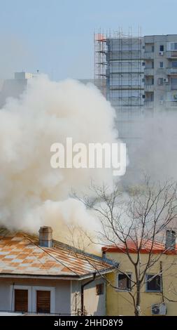 Flammen gehen aus brennendem Haus in der Nachbarschaft. Rauch, der aus dem Dach auf Feuer in der Stadtlandschaft auftaucht. Gefährliche Dämpfe und Smog, die durch eine Explosion aus dem zerstörten Gebäude entstehen Stockfoto