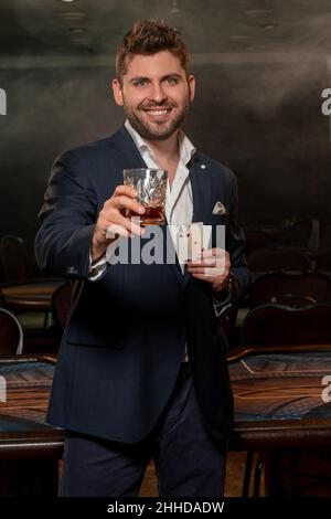 Lächelnder Kerl mit zwei Assen in der Hand, der ein Glas Whiskey zum erfolgreichen Pokerspiel hebt Stockfoto
