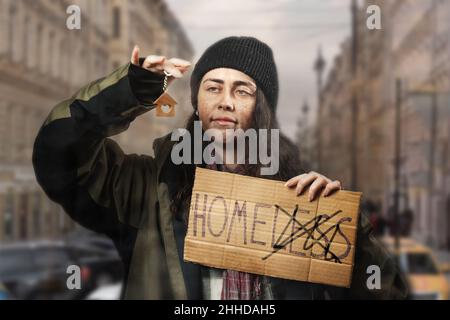 Eine lächelnde Bettlerin hält ein Pappschild mit der Aufschrift Obdachlos und schaut auf einen Schlüsselanhänger in Form eines Hauses. Das Konzept der Unterstützung von VAG Stockfoto