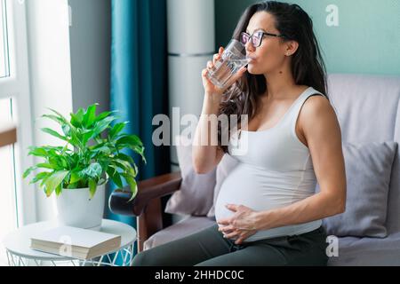 Schwanger junge Frau mit Gläsern trinken Glas Wasser im Wohnzimmer Stockfoto