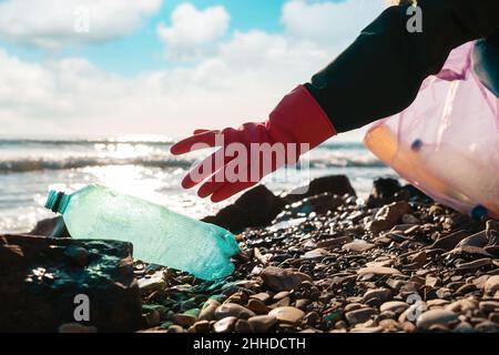 Ein Freiwilliger in Gummihandschuhen greift nach einer schmutzigen grünen Plastikflasche, die am Meeresufer liegt. Nahaufnahme der Hand. Konzept von Save the Planet. Stockfoto