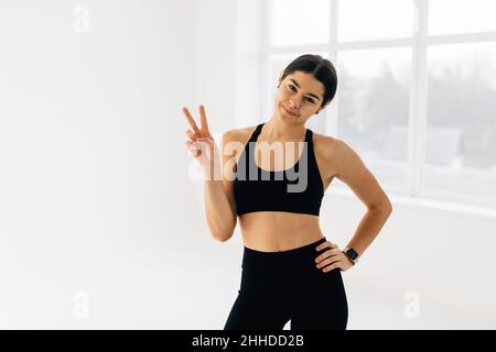 Fit und schlank Fitness-Mädchen zeigt Friedenszeichen und zwinkern, Training im Fitnessstudio, tun Sportübungen in aktivem Ohr, weißer Hintergrund Stockfoto