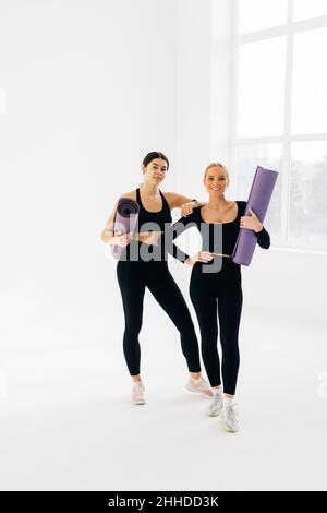 Zwei Freundinnen in Sportswear laufen mit einer Yogamatte zusammen, unterhalten sich und haben Spaß während der Yogapause im Fitnessstudio Stockfoto