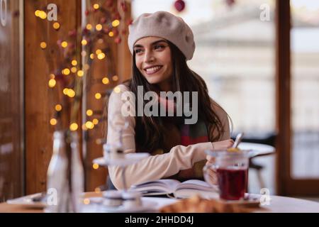 Porträt einer Frau im pariser Stil, die Baskenmütze und Schal trägt, während sie im Café tagernd aufgeht. Stockfoto