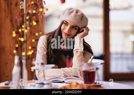 Porträt einer Frau im pariser Stil, die Baskenmütze und Schal trägt, während sie im Café tagernd aufgeht. Stockfoto