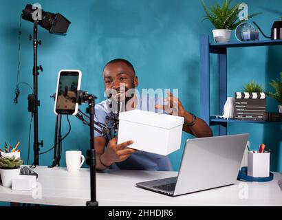 Influencer präsentiert weißes Mockup-Geschenk für Fans, die sich im vlogging-Studio Live-Video-Podcast ansehen. Lächelnder Vlogger hält die Produktbox, die Online-Werbegeschenke vor dem Aufnahmemophone hostet. Stockfoto