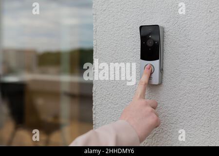 Die Hand der Frau benutzt eine Türklingel an der Wand des Hauses mit einer Überwachungskamera Stockfoto