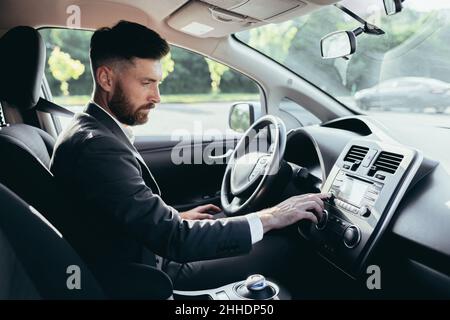 Ein Geschäftsmann, der ein Auto fährt, schaltet die Musik im Auto-Player ein, ein Mann vor einer Reise in einem Geschäftsanzug Stockfoto